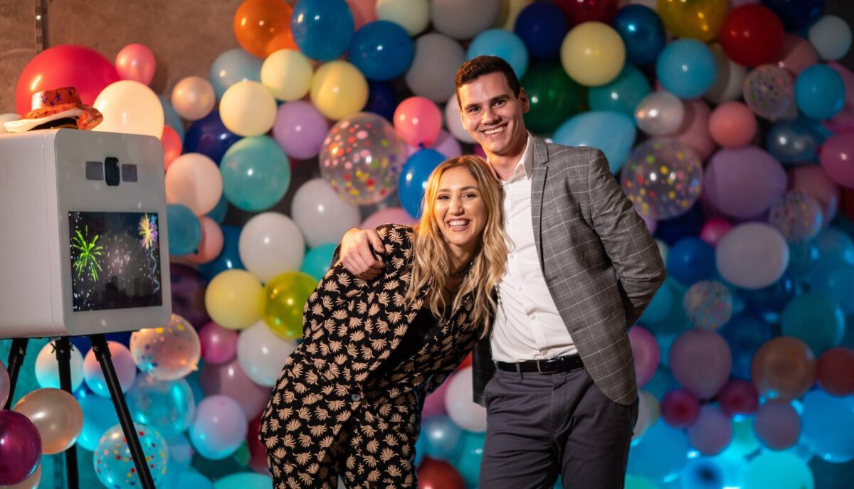 Couple souriant qui pose devant un photobooth et un mur de ballons colorés